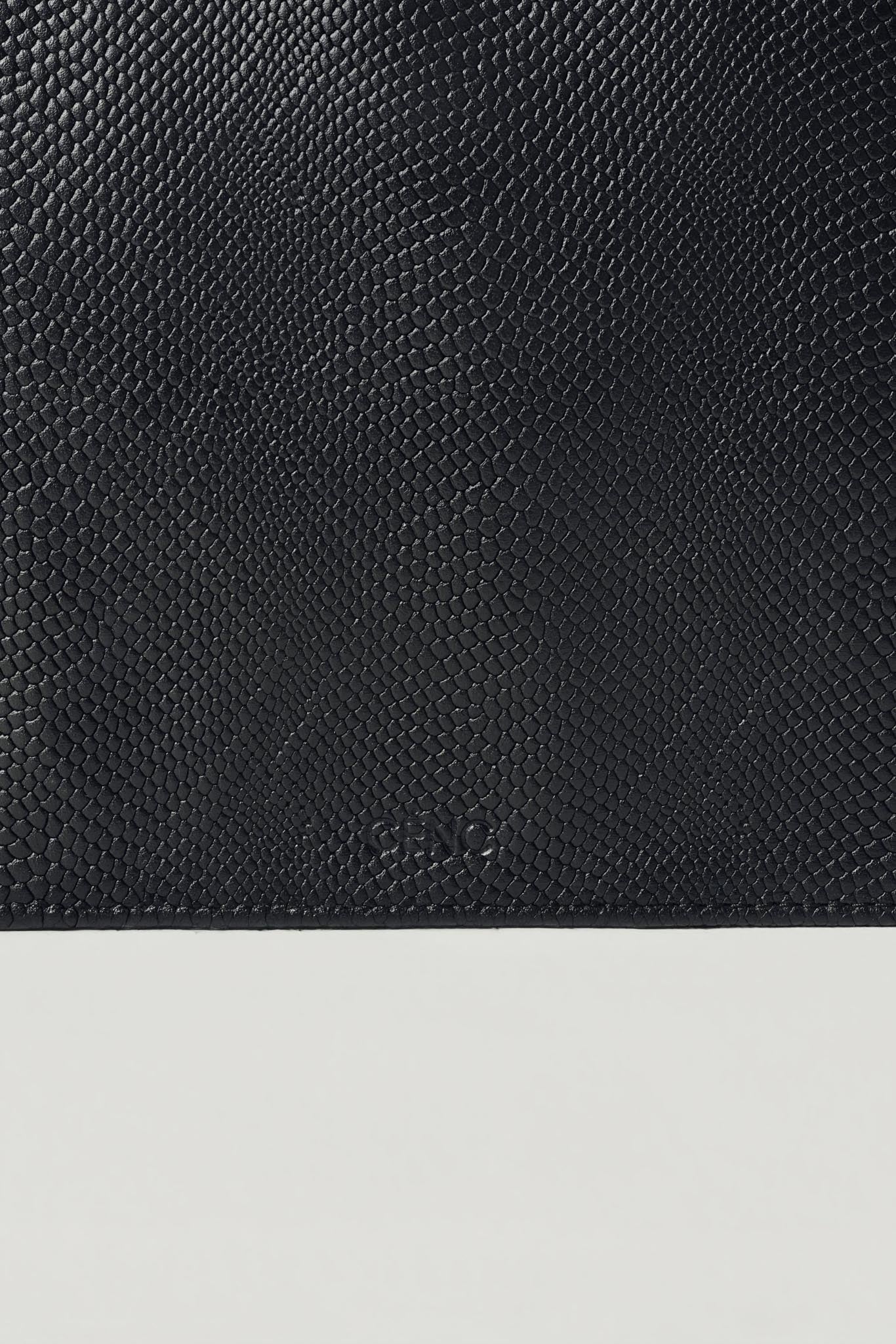 Close-up of a black textured leather bag with a subtle embossed logo, shown against a light gray background.