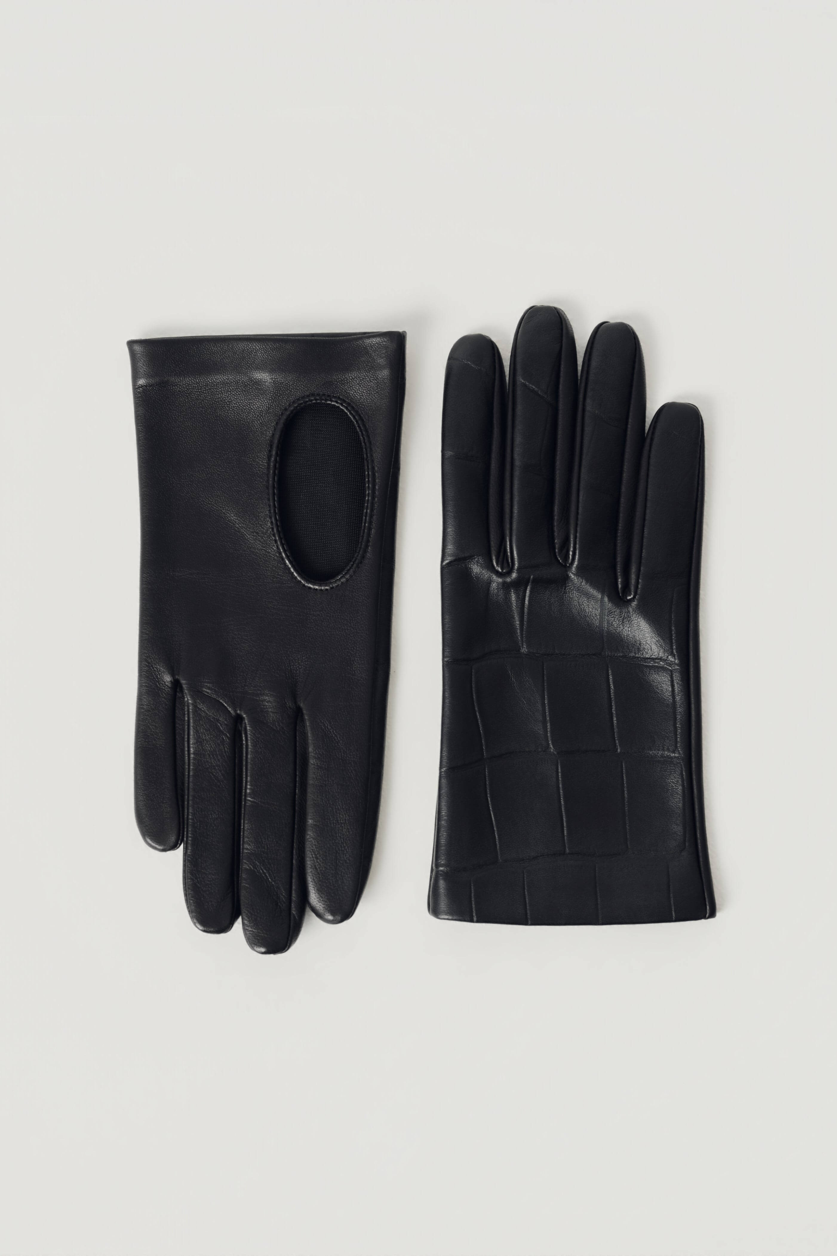 A pair of black leather gloves, one showing the back of the hand with a smooth finish and a circular cutout, and the other showing the palm with a crocodile-embossed texture, laid flat on a light gray background.